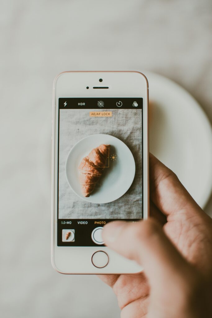 pexels photo 1387444 1387444 A person photographs a croissant on a plate using a smartphone indoors.